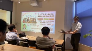 Speaker at a standing desk with a laptop giving a talk, projector screen showing a colorful AI meetup flyer in Japanese; audience seated in a conference room.