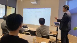 Presenter in a dark suit stands at right giving a training talk, projected screen showing a computer interface on the wall for the seated group.