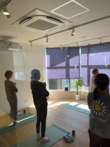 People stand on blue-green yoga mats in a bright office studio, practicing yoga with props nearby (plants, window blinds).