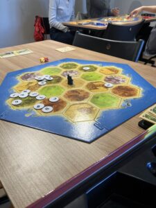 A Settlers of Catan board on a wooden table with hex tiles, dice, and game pieces; players sit around in the background.