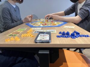 Two people play a tabletop strategy game at a wooden table with an orange-blue hex board and colorful plastic pieces across the board.