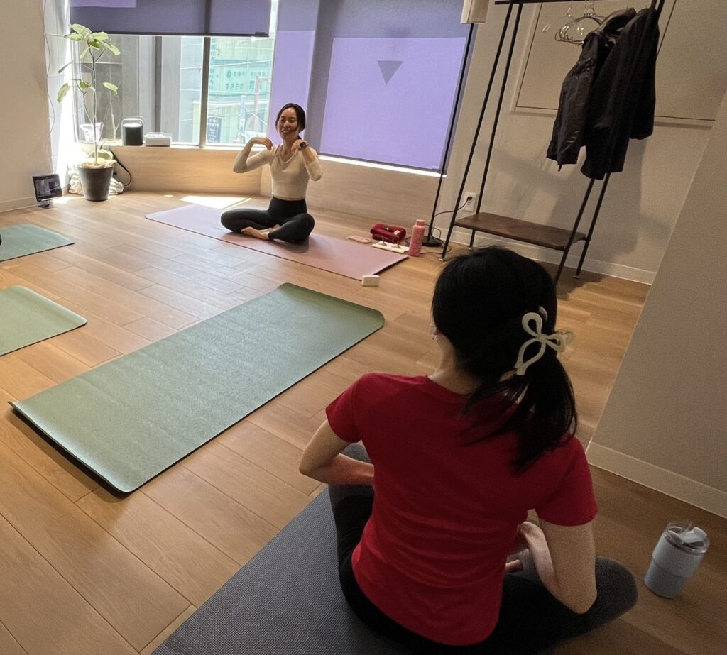 Yoga class in a bright studio: instructor sits on a pink mat demonstrating a pose while a student in a red shirt sits on a blue mat in the foreground, with coats on a rack nearby.