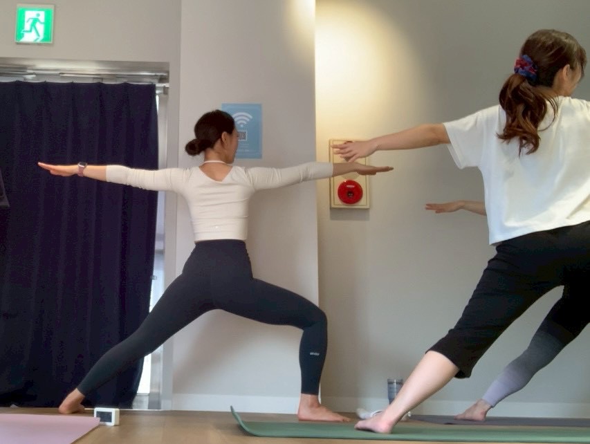 Two women practicing yoga in a studio, arms extended in a wide stance on their mats as they stretch forward and back to either side of the room.