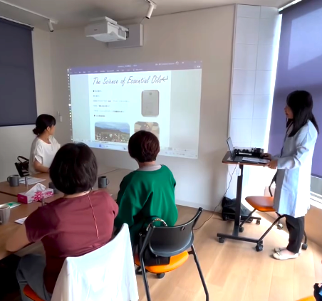 Instructor stands at the front of a bright meeting room, presenting a slide titled "The Science of Essential Oils" to four adults seated around a table; a projector displays the slide on the wall, and a presenter’s cart with equipment sits nearby.