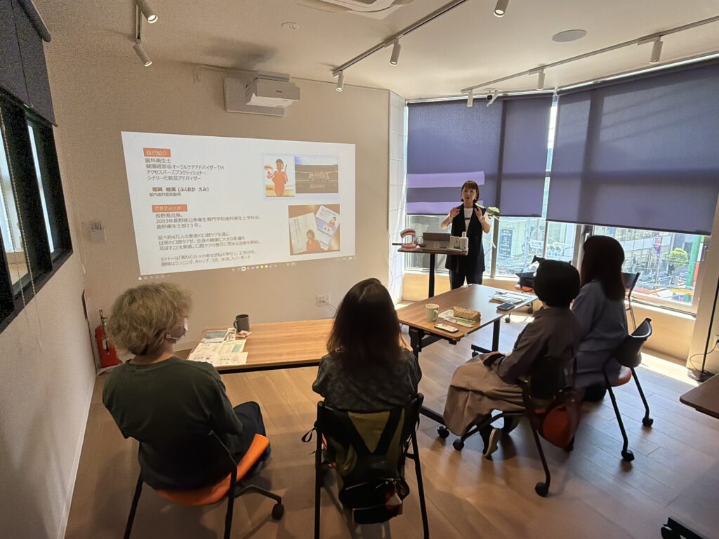 Presenter speaks to a small audience in a bright conference room, projection showing slides with Japanese text and images on the wall/ screen.",