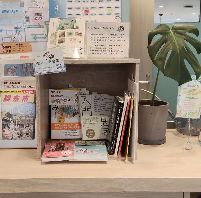 Wooden display shelf with Japanese magazines and books on a desk, a large potted Monstera plant on the right, and posters on the wall behind.