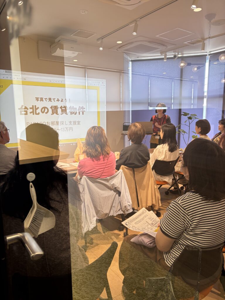 IMG_3118 - Share Nest 仙川 Presenter speaks at a podium in a glass-walled meeting room while a slide on screen mentions Taipei rental properties, with attendees seated and watching.