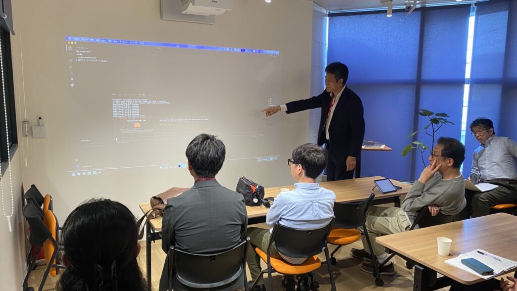 IMG_3124 - Share Nest 仙川 Presenter in a dark suit points at a projected screen on a wall while colleagues sit at tables watching a demonstration in a modern office conference room.