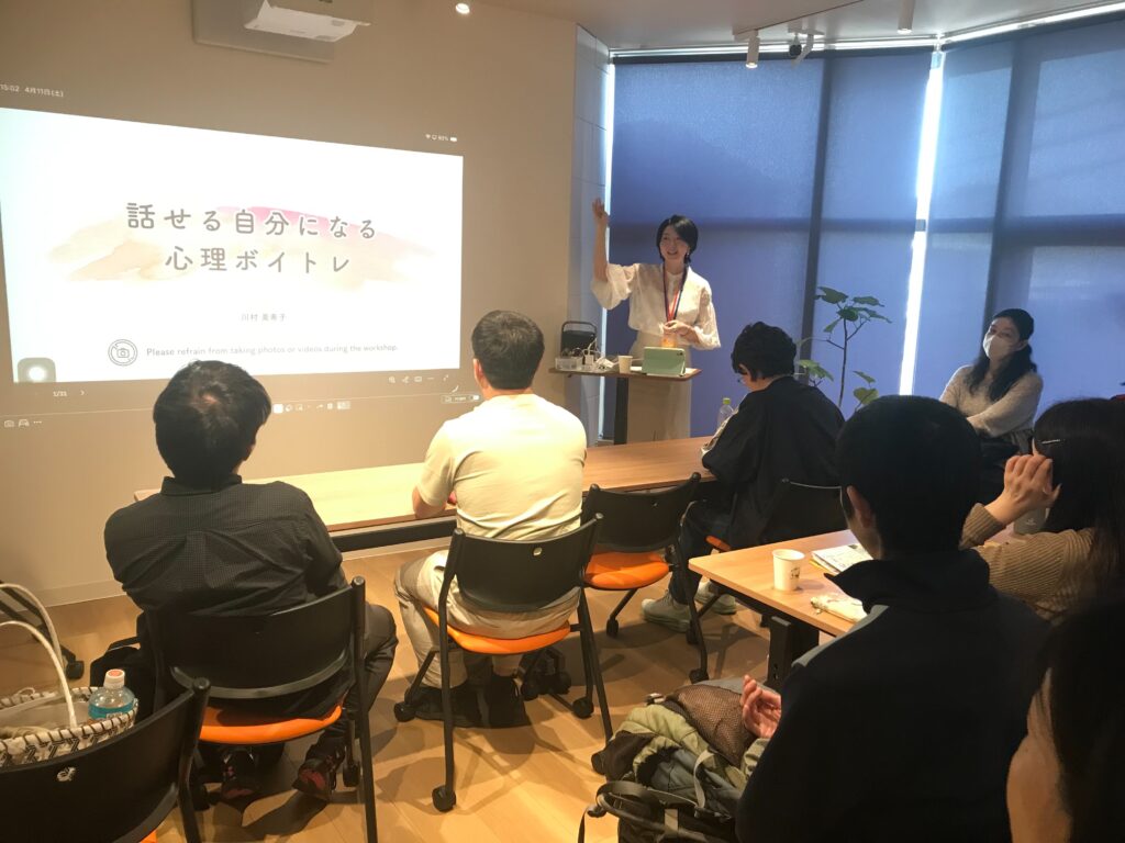IMG_3126 - Share Nest 仙川 Presenter in white blouse standing at the front, raising a hand as a slide is projected onto the wall during a workshop; attendees sit at desks in a room with blue blinds behind them.