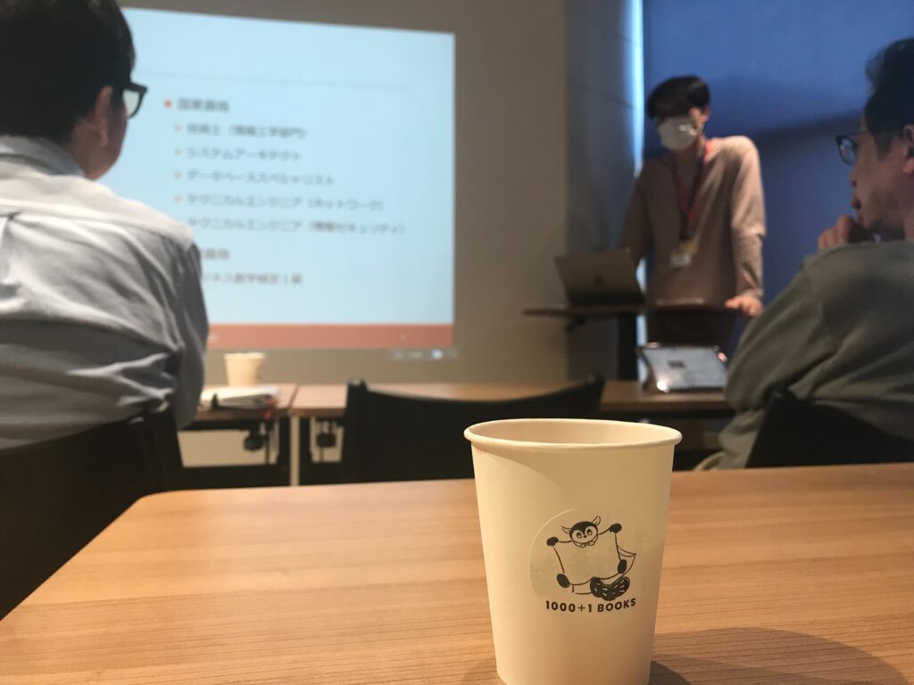 IMG_3128 - Share Nest 仙川 Coffee cup on a wooden table with a projection screen and presenter in a meeting room behind it.