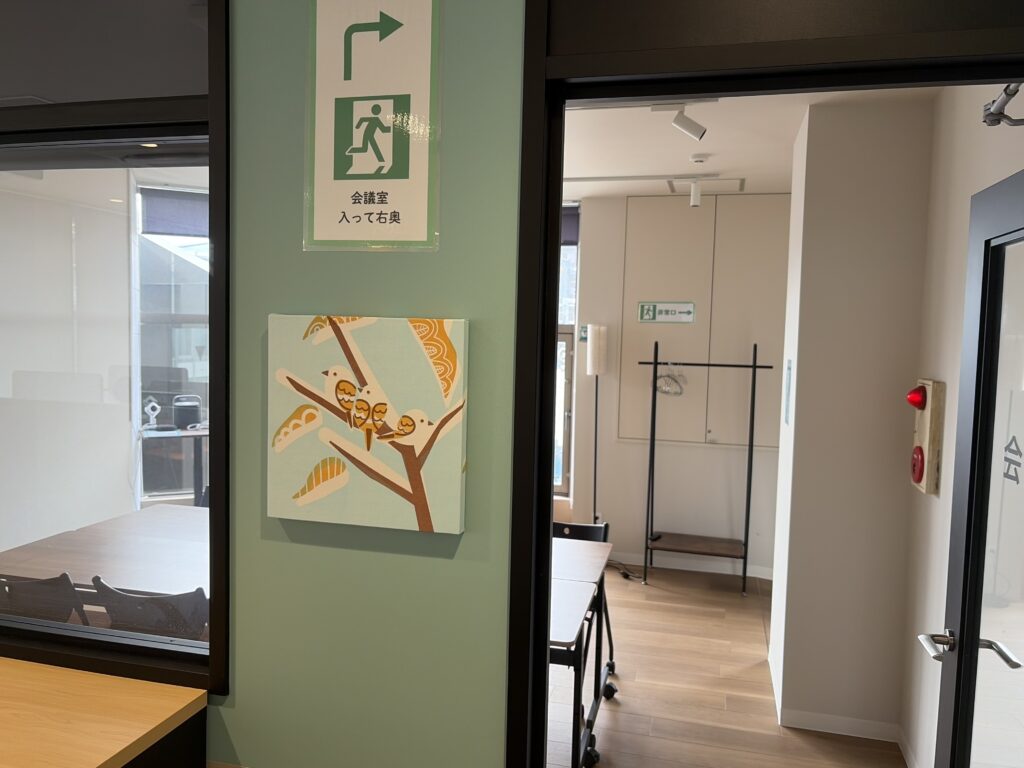 IMG_3727 - Share Nest 仙川 Interior office space with glass walls, a green pillar, and a doorway leading to a conference area; a painting of birds on a branch hangs on the green wall.